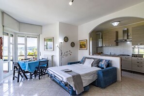 Ein Schlafzimmer mit Bett, einer Couch, einem Esstisch mit Stühlen, einer Küchenzeile und Blick durch ein Fenster.
