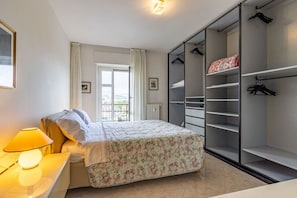 Un dormitorio con cama, lámpara de noche, ventana con cortinas, radiador y armario con perchas.