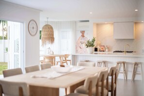 Una cocina moderna con armarios blancos, una zona de comedor con mesas y sillas de madera, y un ventanal con cortinas.