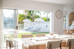Een modern interieur met een eetgedeelte, een grote schuifdeur naar een buitenzitplaats met parasols en uitzicht op groen.