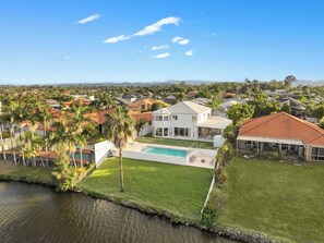 A residential area with a large house featuring a swimming pool, surrounded by palm trees and a body of water. - Whitesands by Aryve | Robina (Robina)