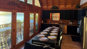 Una cabaña de madera con una mesa de comedor dispuesta para seis personas, una cocina con armarios oscuros y ventanas grandes con vistas a una masa de agua.