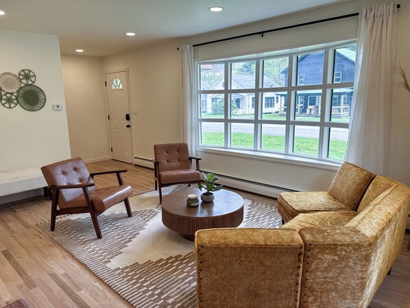 A living room with a sofa, two armchairs, a coffee table, and a large window with a view of houses.