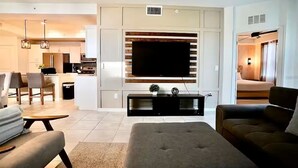 Een moderne woonkamer met een flatscreen televisie, een bank, een salontafel en een eethoek op de achtergrond.