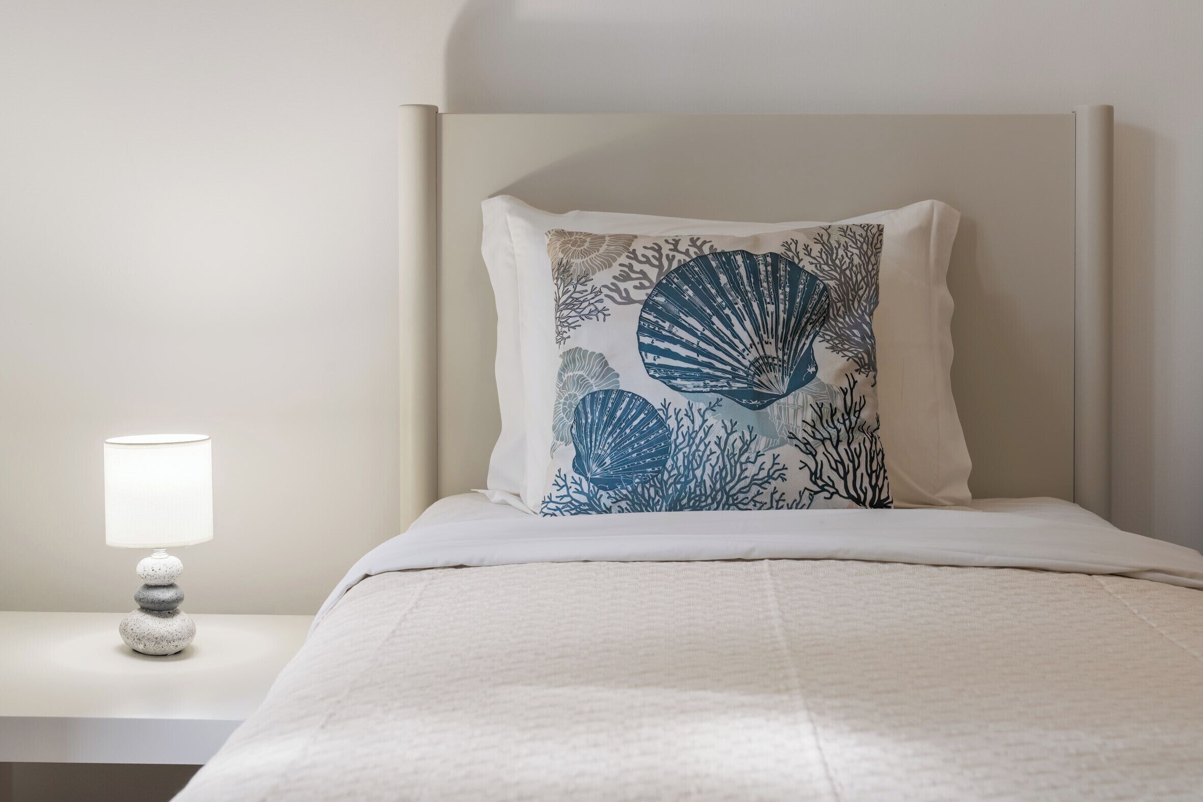 Ein ordentlich bezogenes Bett mit einer weißen Tagesdecke, einem Kissen mit blauem Design aus Meeresschnecken und Korallen, und einer Nachttischlampe auf einem weißen Nachttisch.
