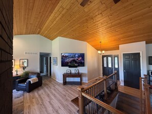 A spacious living room with a wooden ceiling, a television mounted on the wall, and a staircase leading to another level. - Spacious 6-bedroom house in Conifer, CO-Hot tub, Golf Sim, Home gym, Sauna! (Conifer)