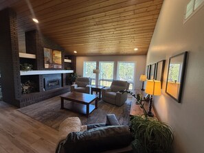 A cozy living room with a fireplace, wooden ceiling, and large windows. - Spacious 6-bedroom house in Conifer, CO-Hot tub, Golf Sim, Home gym, Sauna! (Conifer)