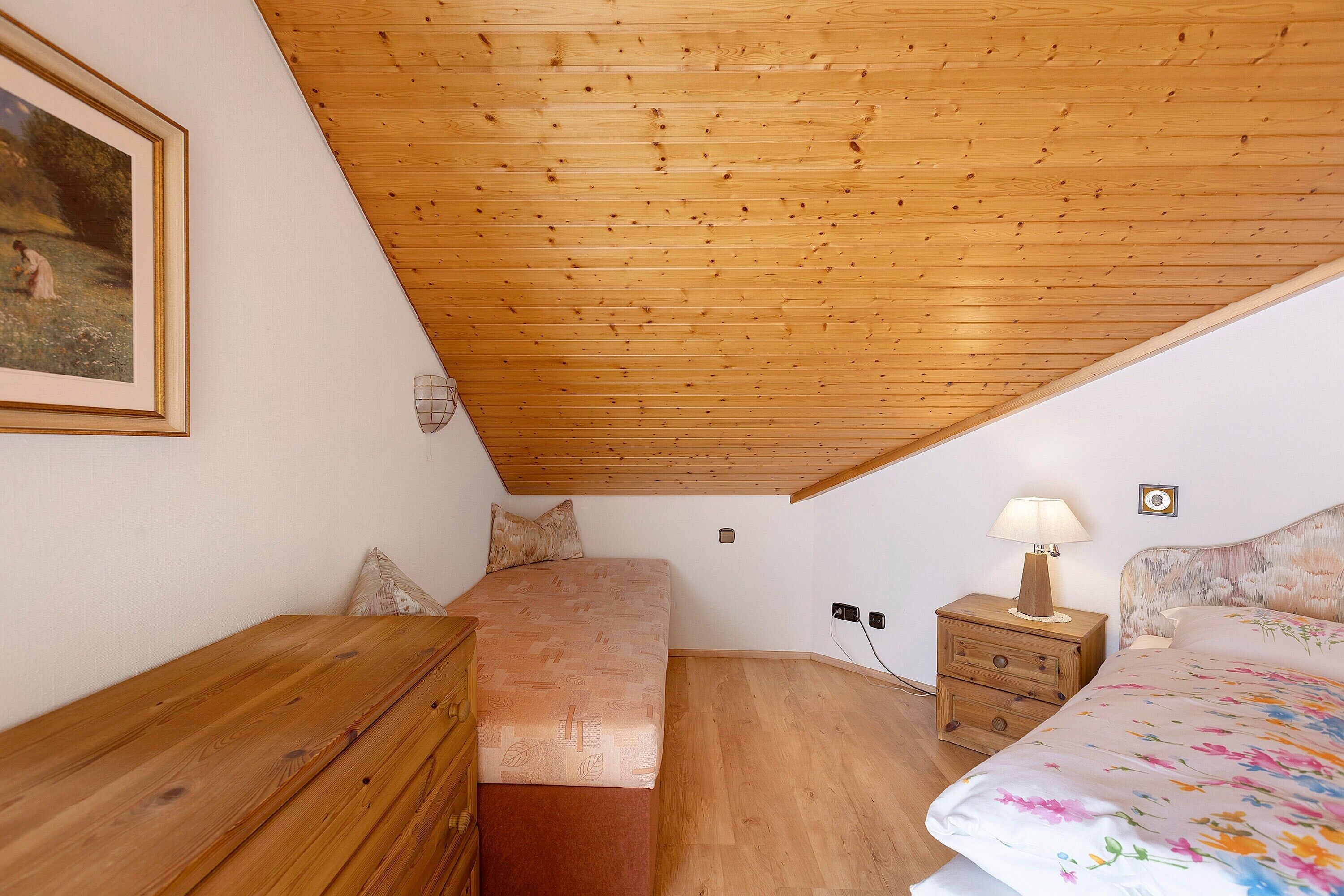Une chambre avec un plafond en pente, des meubles en bois, un lit avec une literie à motifs floraux, une table de chevet avec une lampe et un tableau encadré au mur.