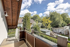 A balcony with a wooden railing, a white wall, and a view of a garden with trees and a building in the background. - Spacious Upstairs Apartment with DreiWelten Card & Balcony near Solemar (Bad Dürrheim)