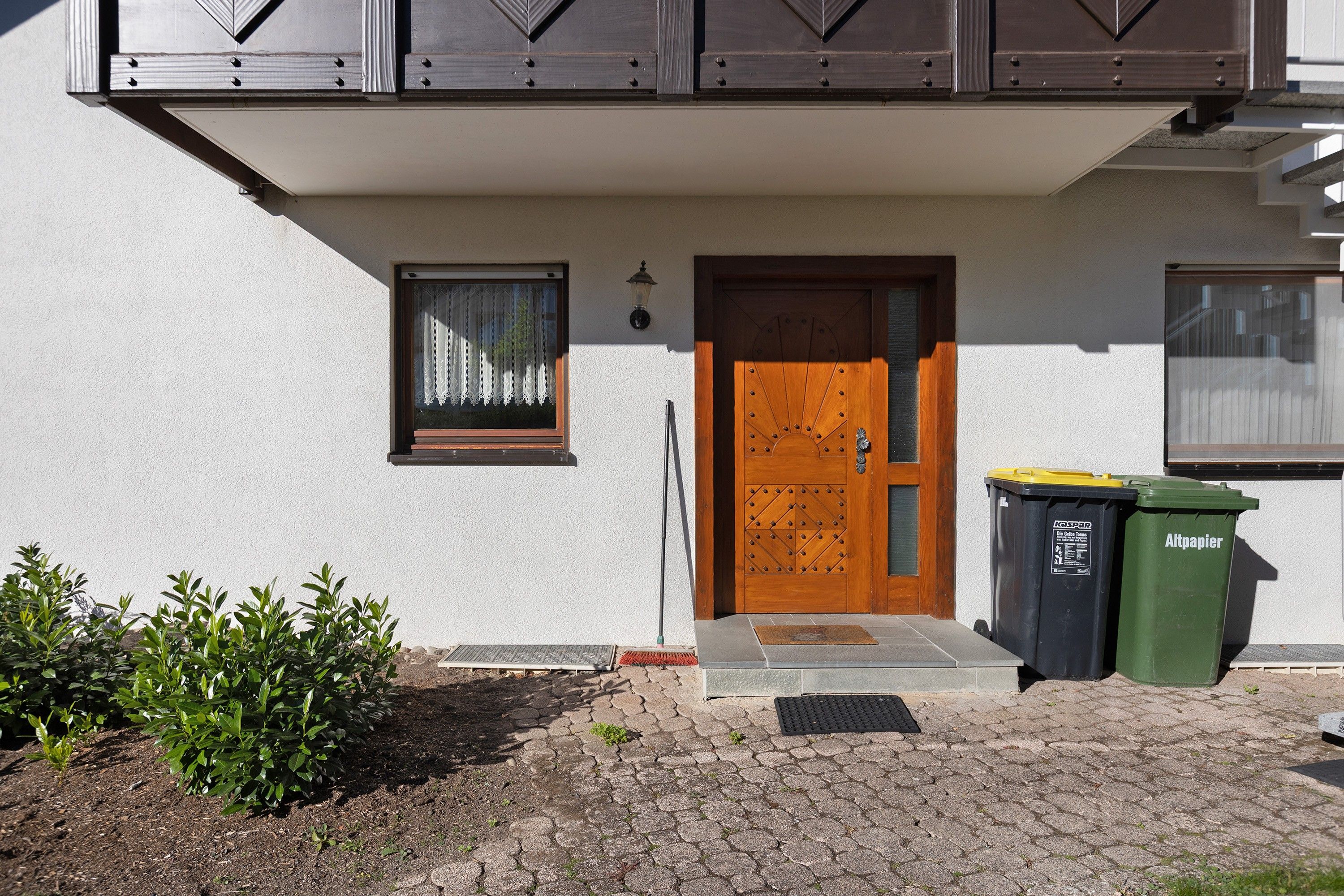 Ein Gebäude mit einer Holztür, einem kleinen Fenster mit Vorhang und zwei Mülltonnen davor.