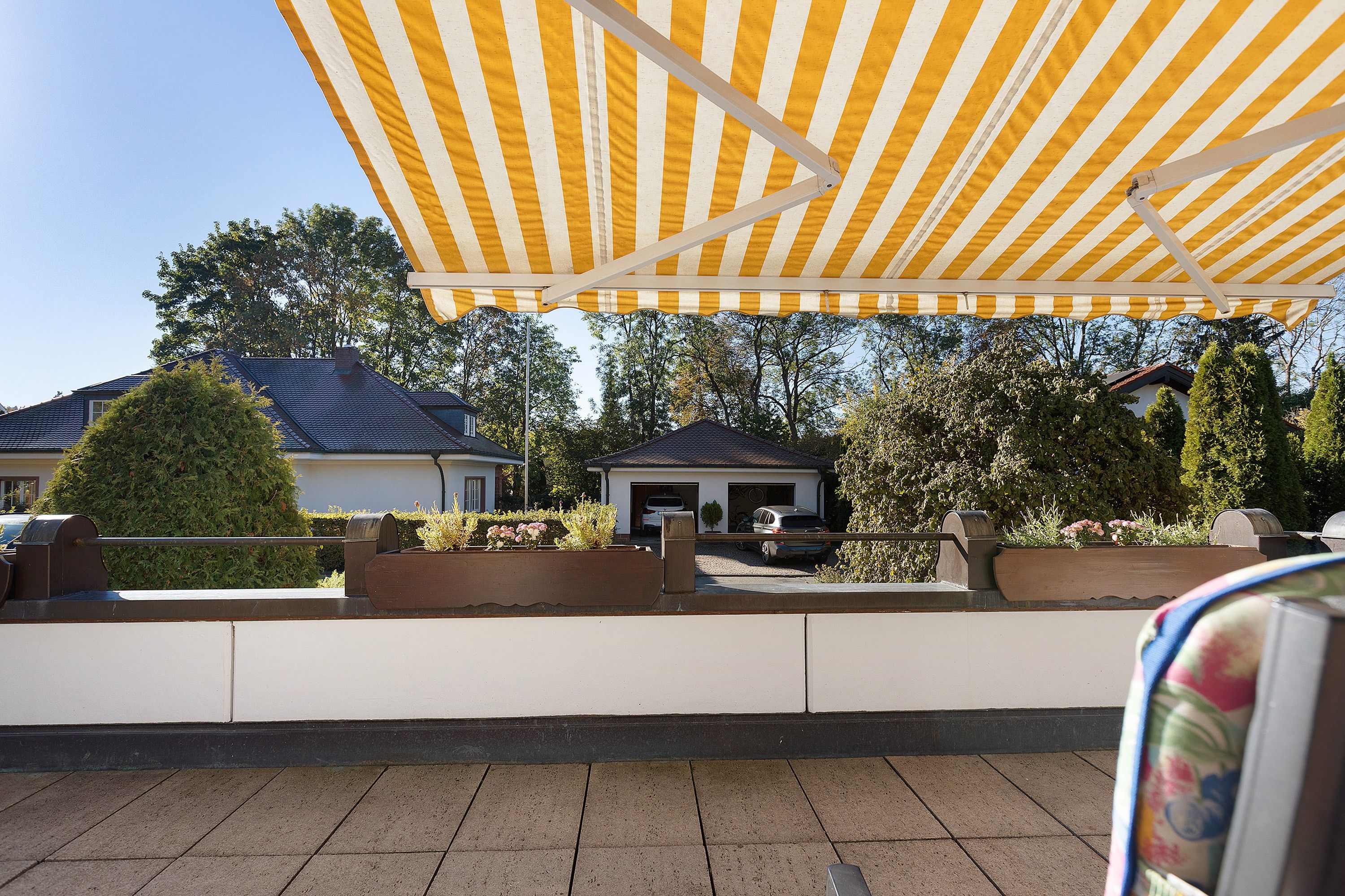 A covered outdoor area with a yellow-striped awning, a paved patio, and a view of residential buildings and trees.