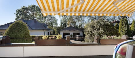 Een overdekt terras met een geel gestreepte luifel, een verhard terras en uitzicht op woongebouwen en bomen.