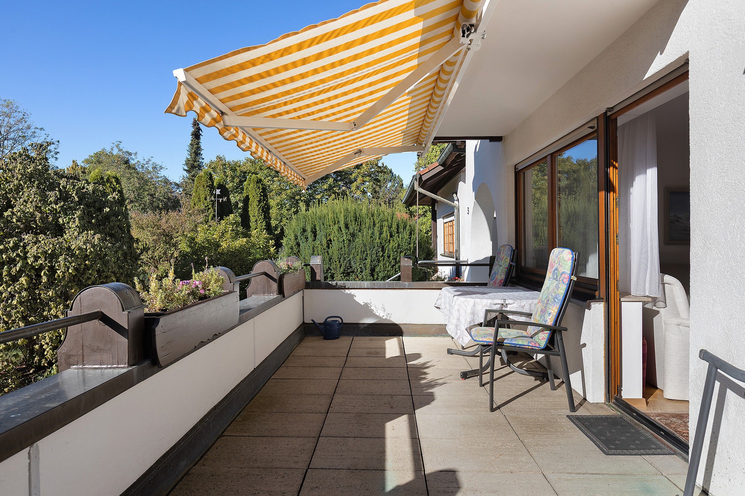 Ein Balkon mit Tisch und Stühlen, einem gelb gestreiften Sonnenschirm und Blick auf Bäume und Himmel.