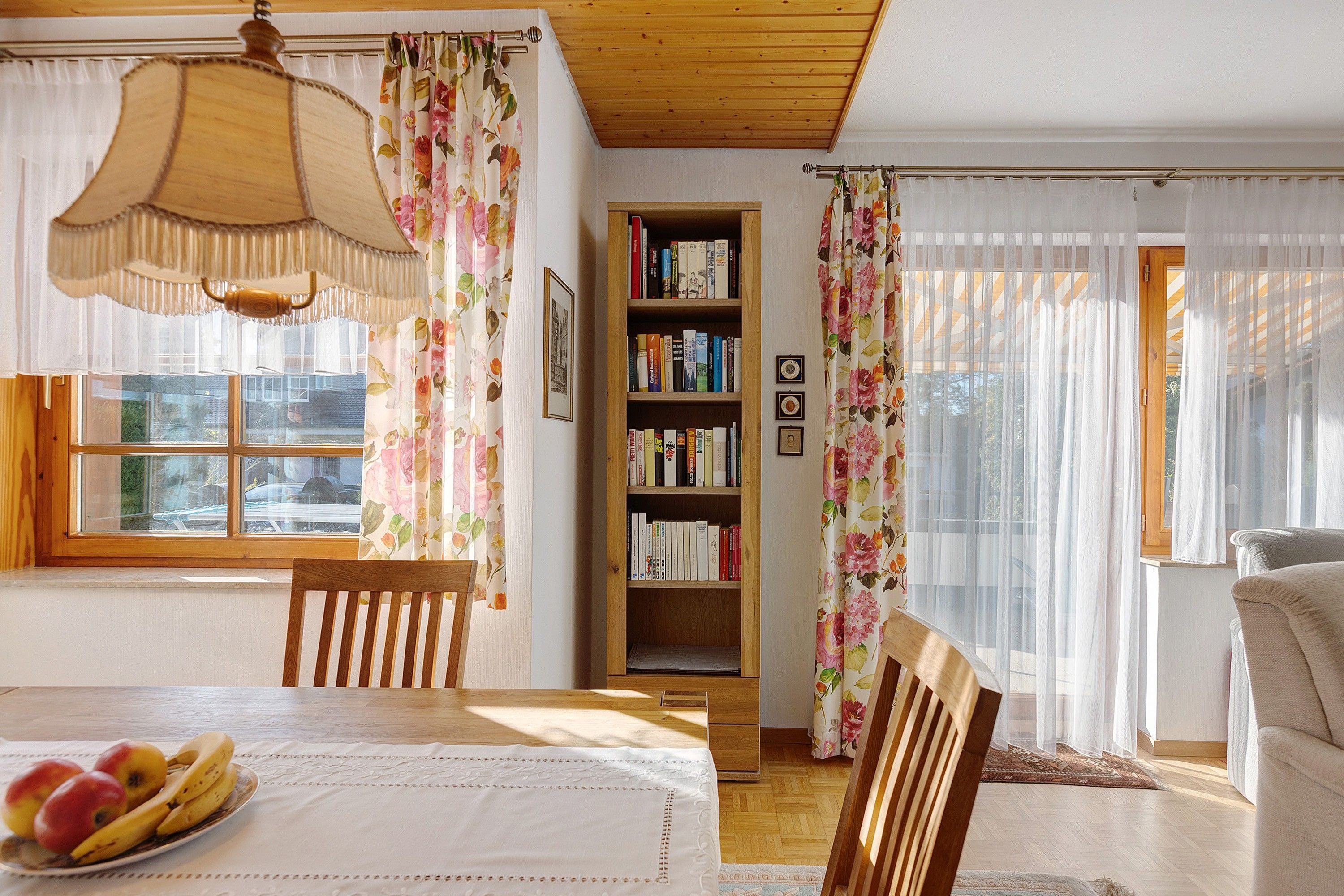 Ein gemütliches Wohnzimmer mit Holzbalkendecke, einem Esstisch mit Obst, einem Bücherregal und blumigen Vorhängen.