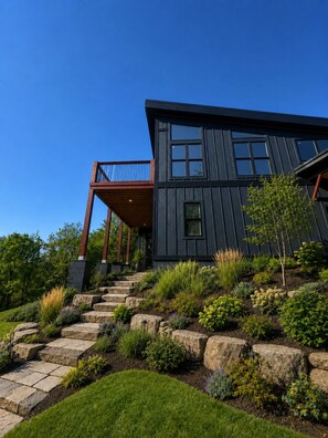 A modern two-story house with a dark exterior, a wooden deck, and a landscaped garden with various plants and a stone pathway leading to the entrance.