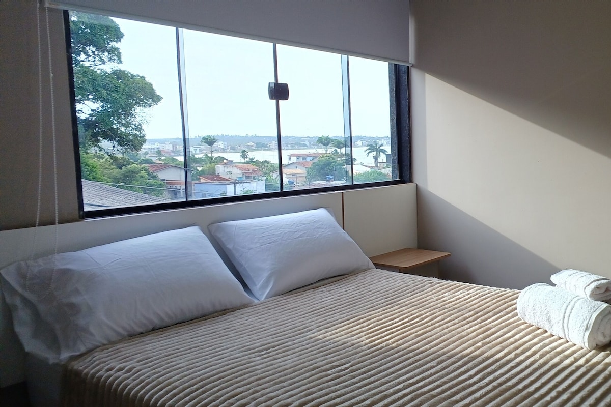 Habitación de hotel con cama, ventana con vista a edificios, una mesita de noche pequeña y una toalla sobre la cama.