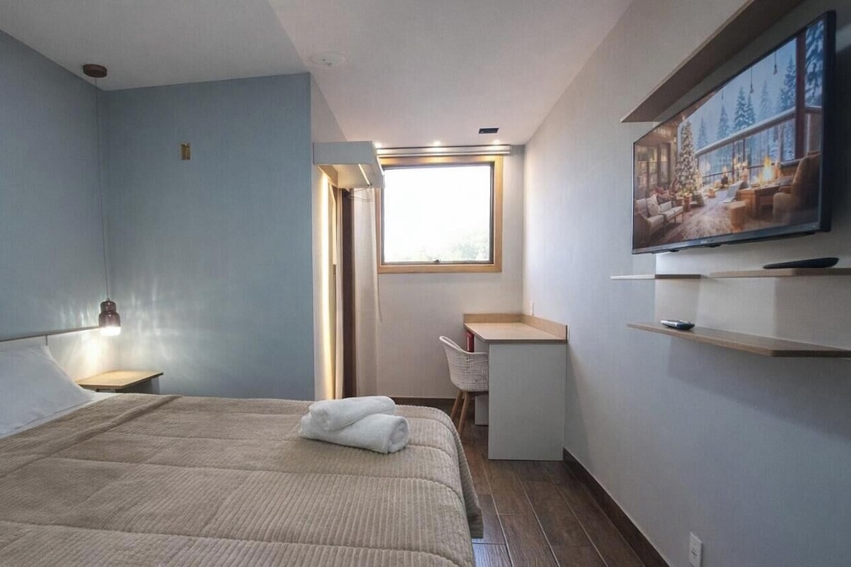 A bedroom with a bed, a desk, a chair, a window, and a television.