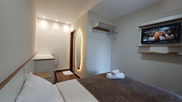 Quarto de hotel com uma cama, uma televisão, um espelho e uma pequena mesa com uma toalha.