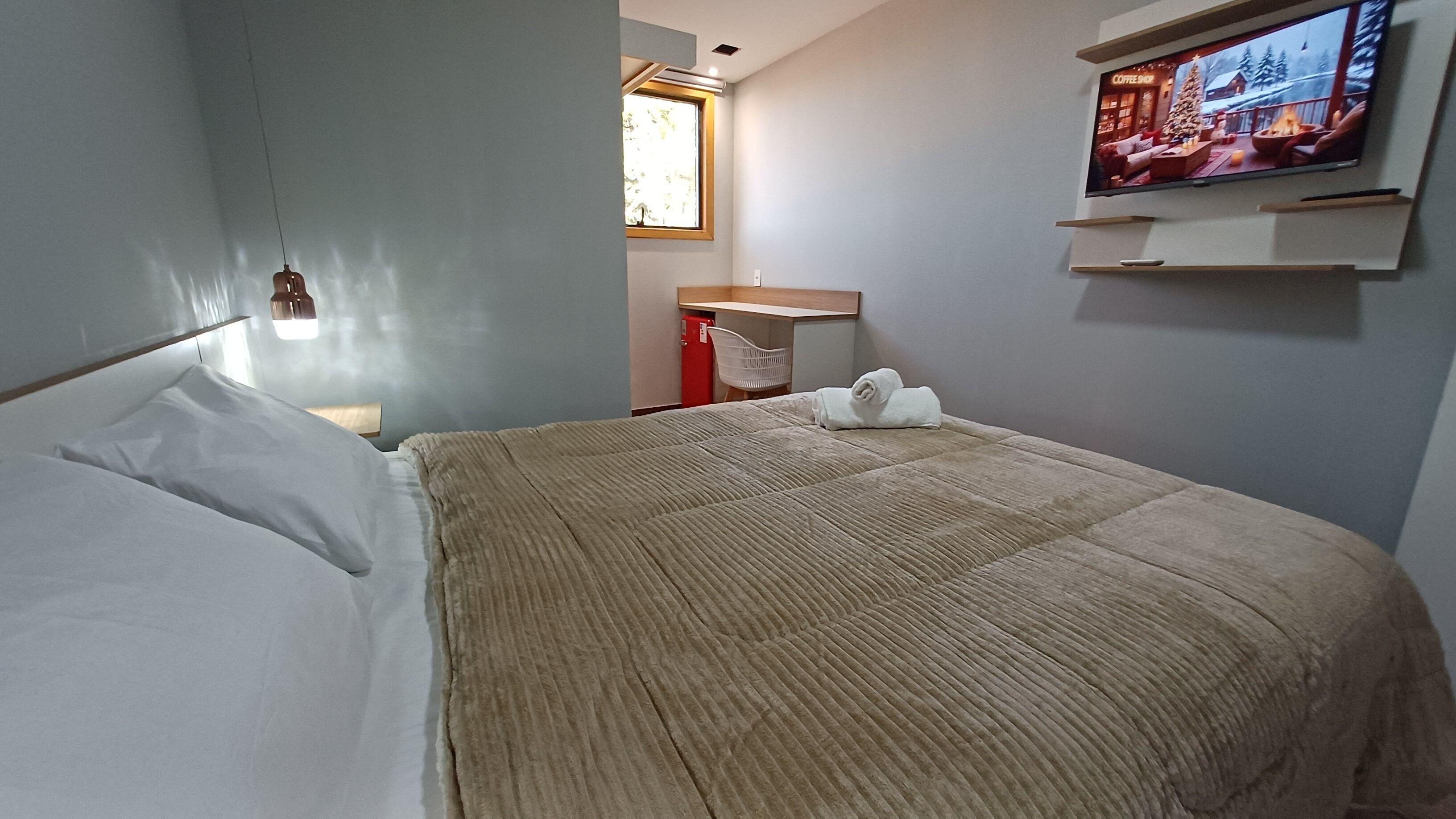 Una camera d'albergo con un letto, una TV a muro, una finestra e una scrivania con una sedia.