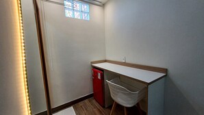 Um quarto pequeno e estreito com uma escrivaninha branca, um eletrodoméstico vermelho e uma cadeira branca.