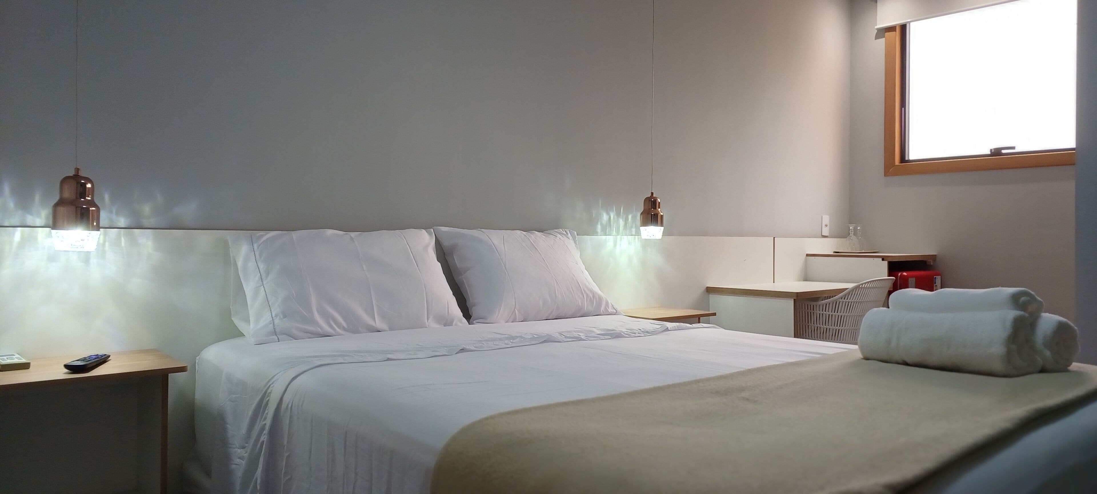A hotel room with a bed, bedside tables, a desk, a chair, a window, and a wall-mounted lamp.