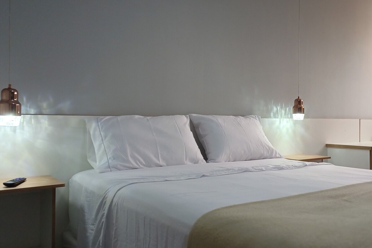 Habitación de hotel con cama, mesitas de noche, escritorio, silla, ventana y lámpara de pared.