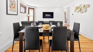 Uma sala de jantar com uma mesa posta para seis pessoas, uma televisão de tela plana na parede e um objeto decorativo na parede.
