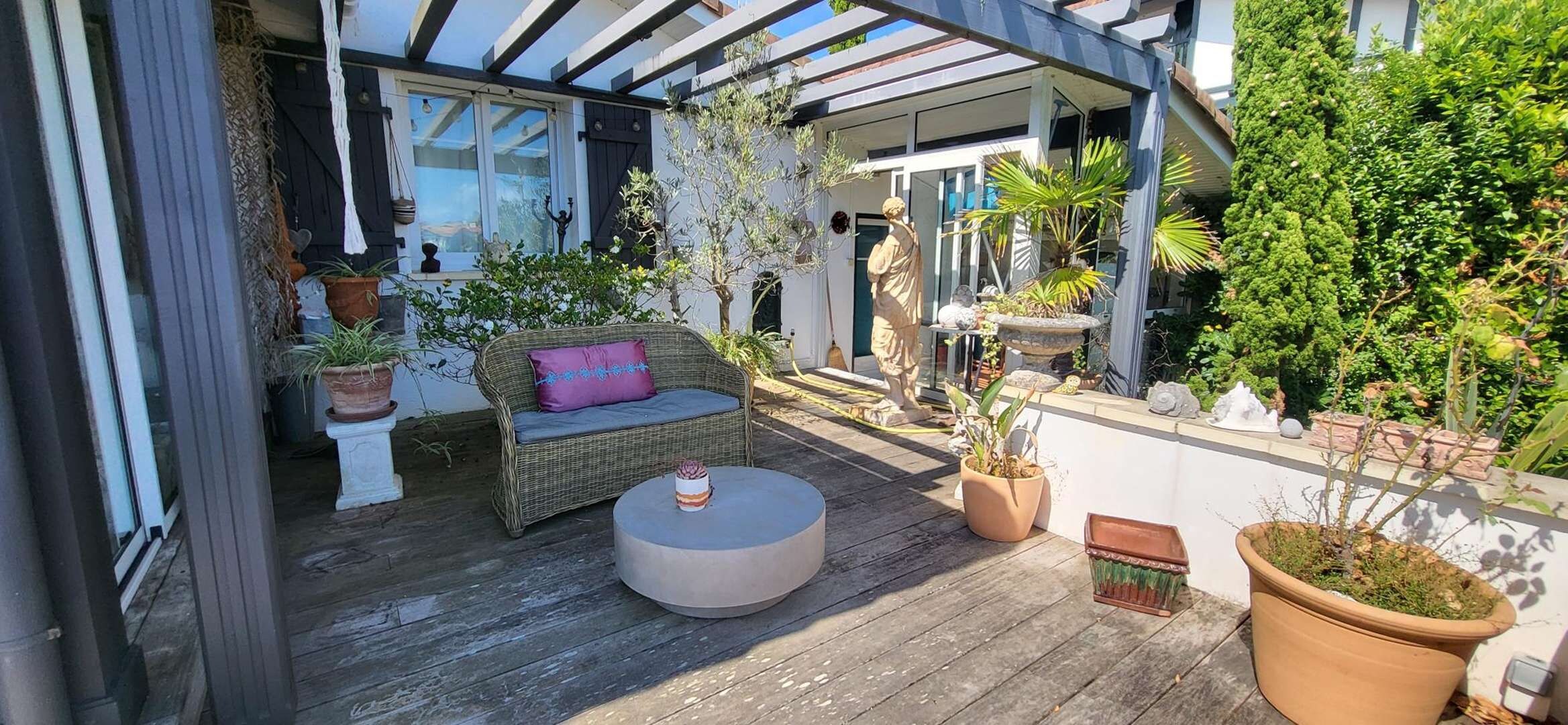 A covered outdoor patio with a wicker sofa, a round table, and various potted plants.