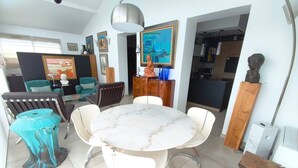 Een moderne woonkamer met een marmeren tafel, witte stoelen, een turquoise accentstoel, een houten kast en een schilderij aan de muur.