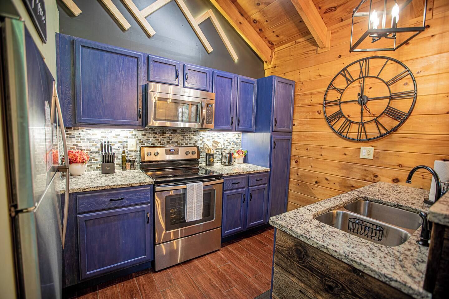 Une cuisine avec des meubles de couleur bleu foncé, des appareils électroménagers en acier inoxydable, un plan de travail en granit, un plancher en bois et une grande horloge murale.