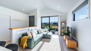 Ein modernes Wohnzimmer mit einem Ecksofa, einem Holz-TV-Schrank und einem großen Fenster mit Blick ins Freie.