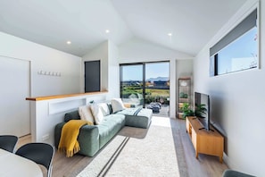 A modern living room with a sectional sofa, a wooden TV stand, and a large window offering a view of the outdoors. - The Snug at Glen Dene (Wānaka)