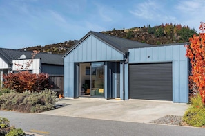 A modern building with a garage door, a driveway, and a hillside in the background. - The Snug at Glen Dene (Wānaka)