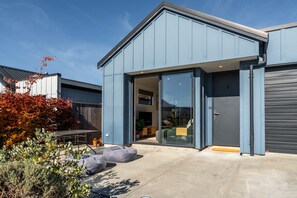 A modern building with a garage door, a patio with seating, and a large glass door leading to an interior space. - The Snug at Glen Dene (Wānaka)