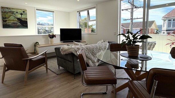 Ein modernes Wohnzimmer mit Glastisch, Ledersesseln, einer Couch, einem Fernseher und großen Fenstern mit Blick auf die Nachbarschaft.