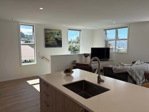 A modern kitchen with a sink, a dining area with a table and chairs, a flat-screen TV, and large windows with a view of the landscape.