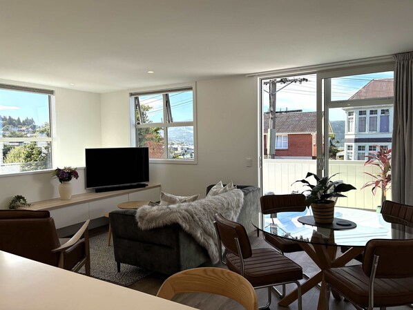 A modern living room with a flat-screen TV, comfortable seating, a glass table, and large windows offering a view of the neighborhood.