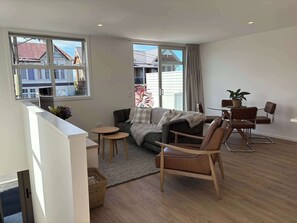 A modern living room with a sofa, a small round table, a dining table with chairs, and a balcony with a view of buildings.