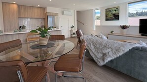 Eine moderne Küche mit Holzschränken, ein Essbereich mit einem Glastisch und Stühlen, eine gemütliche Wohnstube mit einer Couch und einem Fernseher, sowie ein großes Fenster mit Blick.