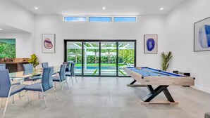 Ein modernes Zimmer mit Pooltisch, Essbereich und großen Fenstern mit Blick auf einen Garten.