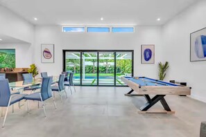 Ein modernes Zimmer mit Pooltisch, Essbereich und großen Fenstern mit Blick auf einen Garten.