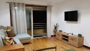 Una sala de estar moderna con sofá, una mesita de centro de cristal, un mueble de televisión de madera y un gran ventanal corredizo que da a un balcón.