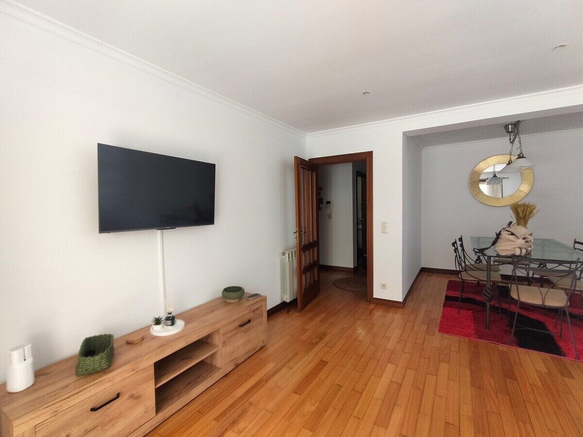 Une pièce dotée d’un téléviseur à écran plat fixé au mur, d’un meuble de rangement en bois, d’une table à manger avec des chaises et d’un tapis rouge.