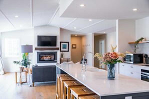 A modern kitchen with a central island, wooden stools, a TV mounted above the fireplace, and a microwave. - Farrant Drive Hideaway with Mountain Views (Wānaka)