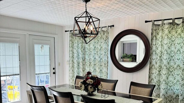 En spiseplads med et bord, stole, en hængende lampe, et spejl og mønstrede gardiner.