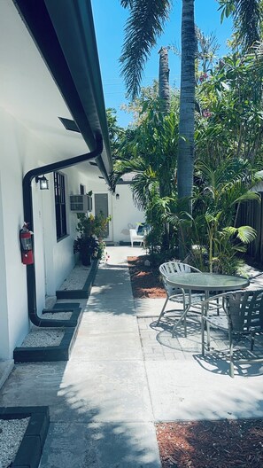 Un patio techado al aire libre con una mesa y sillas, un extintor y un sendero que conduce a la entrada de un edificio.