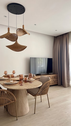 A modern dining area with a round table, wicker chairs, and a flat-screen TV in the background.