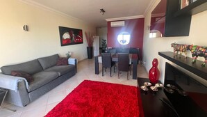 Una sala de estar con un sofá gris, un tapete rojo, una zona de comedor con mesa y sillas, y un mueble bajo para televisión con figuritas.