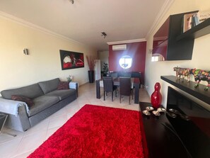 Ein Wohnzimmer mit grauem Sofa, einem roten Teppich, einem Essbereich mit Tisch und Stühlen sowie einer TV-Konsole mit Figuren.