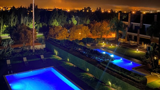 Una vista nocturna de un resort con piscinas iluminadas, una palmera y un paisaje urbano distante.
